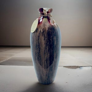May include: A tall, decorative vase with a unique, elongated shape. The vase features a blend of brown, blue, and white vertical streaks, creating a textured appearance. The top is a darker maroon color, tied with twine, and includes a small tag. The vase is approximately 18 inches tall.