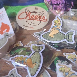 Może przedstawiać: Kolekcja naklejek i naszywek. Jedna naklejka przedstawia słowo "Cheers" z ilustracją osła. Kilka naszywek przedstawia kobietę w bikini jadącą na bombie. Widoczne są inne naszywki i naklejki.