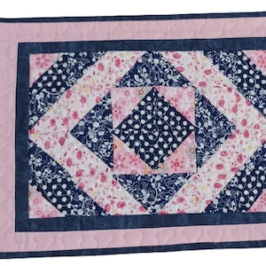 Könnte beinhalten: Ein rechteckiger, gesteppter Tischset mit einem rosa Rand und einem zentralen Design mit marineblauen Punkten und floralen Mustern. Ein Abschnitt mit blau-weißen Streifen befindet sich auf der rechten Seite. Das Tischset ist ca. 30 cm breit und 46 cm lang.