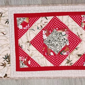 Peut inclure: Un set de table matelassé avec un motif central rouge en forme de carré dans un carré avec des pois blancs et des motifs floraux. La bordure est rouge et les côtés présentent un tissu crème avec des grues et du feuillage. Mesure environ 30 cm de long et 20 cm de large.