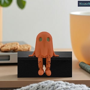 May include: A terracotta ghost figure with jointed limbs sits on a black box with a textured surface. The ghost has two green eye-holes. The background includes a mug, biscuits, and a pen. The text "HouseNHold" is in the upper right corner.