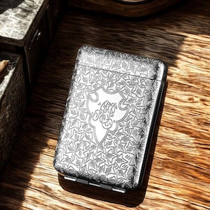 May include: A silver cigarette case with an engraved floral design. The case has a central crest with initials. The rectangular case has rounded edges and a hinged closure. It is displayed on a wooden surface.