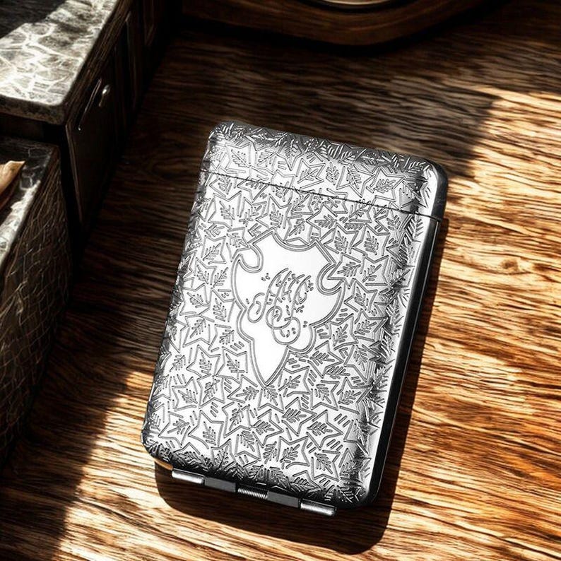 May include: A silver cigarette case with an engraved floral design. The case has a central crest with initials. The rectangular case has rounded edges and a hinged closure. It is displayed on a wooden surface.
