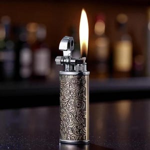 May include: A vintage-style lighter with an ornate floral pattern etched into its brass-colored body. The lighter is lit, with a tall, yellow flame extending upwards. The top portion is silver-colored, and the background is blurred.