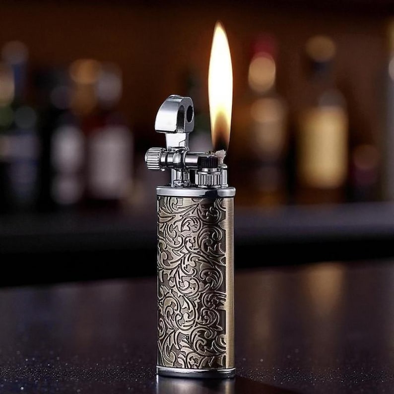 May include: A vintage-style lighter with an ornate floral pattern etched into its brass-colored body. The lighter is lit, with a tall, yellow flame extending upwards. The top portion is silver-colored, and the background is blurred.