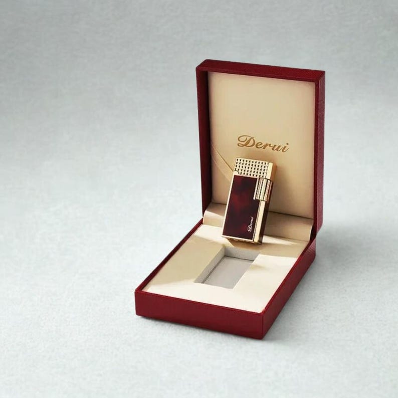 May include: A red and gold lighter with the brand name "Derui" is displayed inside a red box with a cream-colored interior. The lighter has a rectangular shape with gold accents and a dark red body. The box is open, showcasing the lighter.