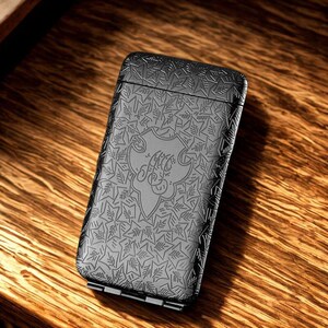 May include: A black metal lighter with an embossed leaf pattern and a decorative emblem. The rectangular lighter has a hinged base and rests on a wooden surface. The design is detailed, giving a textured appearance.