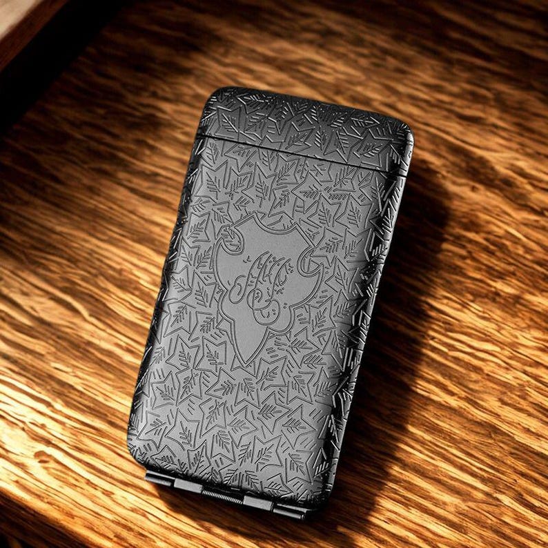 May include: A black metal lighter with an embossed leaf pattern and a decorative emblem. The rectangular lighter has a hinged base and rests on a wooden surface. The design is detailed, giving a textured appearance.