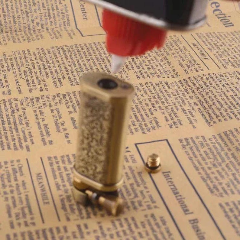 May include: A vintage brass lighter with an ornate, textured design. The lighter is rectangular with rounded edges and a small, separate screw-on piece. It sits on a newspaper background.