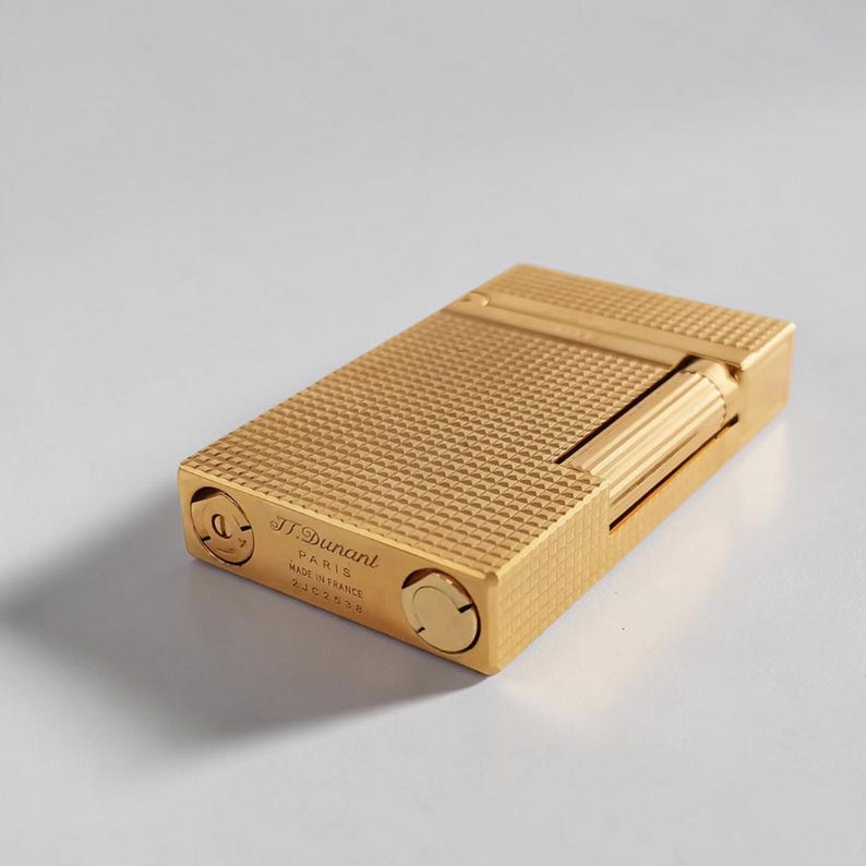 Puede incluir: Un encendedor rectangular dorado con una superficie texturizada con patr&oacute;n de diamantes. El encendedor tiene una tapa con bisagras y un rodillo cil&iacute;ndrico. El texto "J. Dupont PARIS MADE IN FRANCE" es visible.