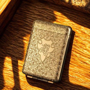 May include: A gold-coloured metal cigarette case with an ornate, engraved floral pattern. The case features a central emblem with stylized script. The case is rectangular with rounded edges and a hinged opening. The case is resting on a wooden surface.