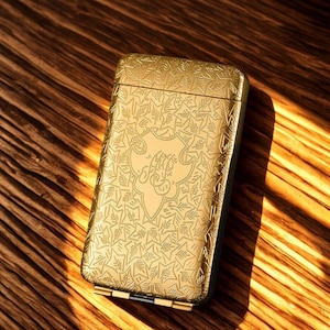 May include: A gold-coloured rectangular case with an ornate leaf and star pattern. The case has a hinged lid and a decorative emblem in the centre. The case is resting on a wooden surface.