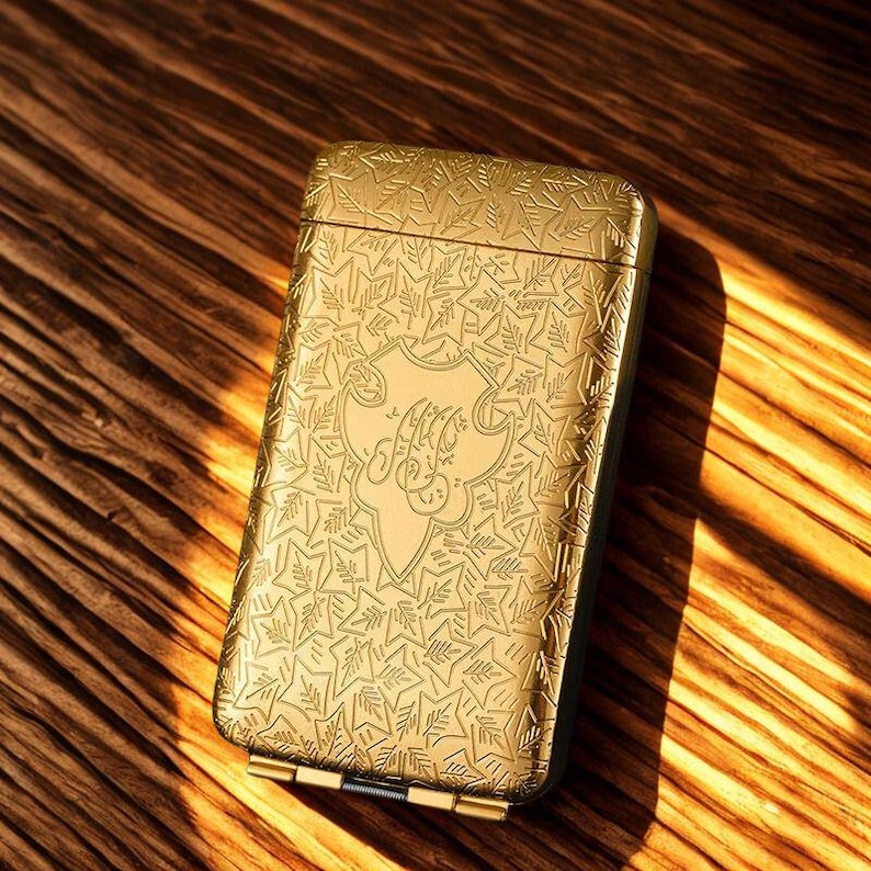 May include: A gold-coloured rectangular case with an ornate leaf and star pattern. The case has a hinged lid and a decorative emblem in the centre. The case is resting on a wooden surface.
