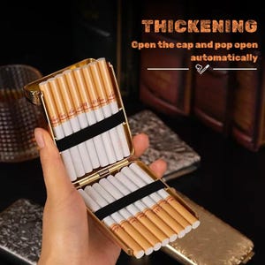 May include: A gold cigarette case, open to show cigarettes arranged in rows. The case has a hinged lid and a black elastic band. The image includes the text "THICKENING" and "Open the cap and pop open automatically."