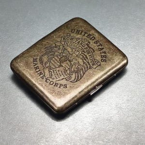 May include: An antique brass-coloured cigarette case. The case is engraved with the words "UNITED STATES MARINE CORPS" and "KOH TANG SURVIVOR" around a detailed emblem. It has a hinged opening and a slightly worn appearance.