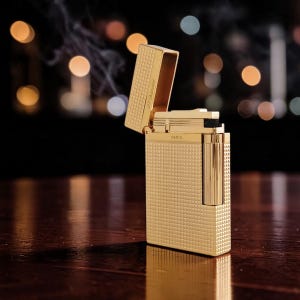 Puede incluir: Un encendedor dorado con una superficie texturizada con patr&oacute;n de diamantes. La tapa est&aacute; abierta, revelando el mecanismo interno. El encendedor est&aacute; marcado "S.T. Dupont Paris". El humo se eleva desde la parte superior, sugiriendo un uso reciente.