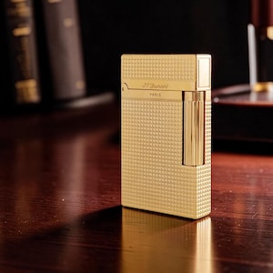 Puede incluir: Un encendedor dorado con una superficie texturizada con patr&oacute;n de diamantes. El encendedor tiene forma rectangular con una tapa con bisagras y una secci&oacute;n vertical y acanalada en el lateral. El texto "J.T. Durant PARIS" es visible.