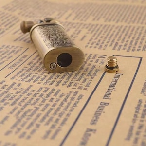 May include: A vintage-style brass lighter with intricate floral engravings. The lighter has a rectangular body with rounded edges and a large circular opening. A small, separate screw is also visible.