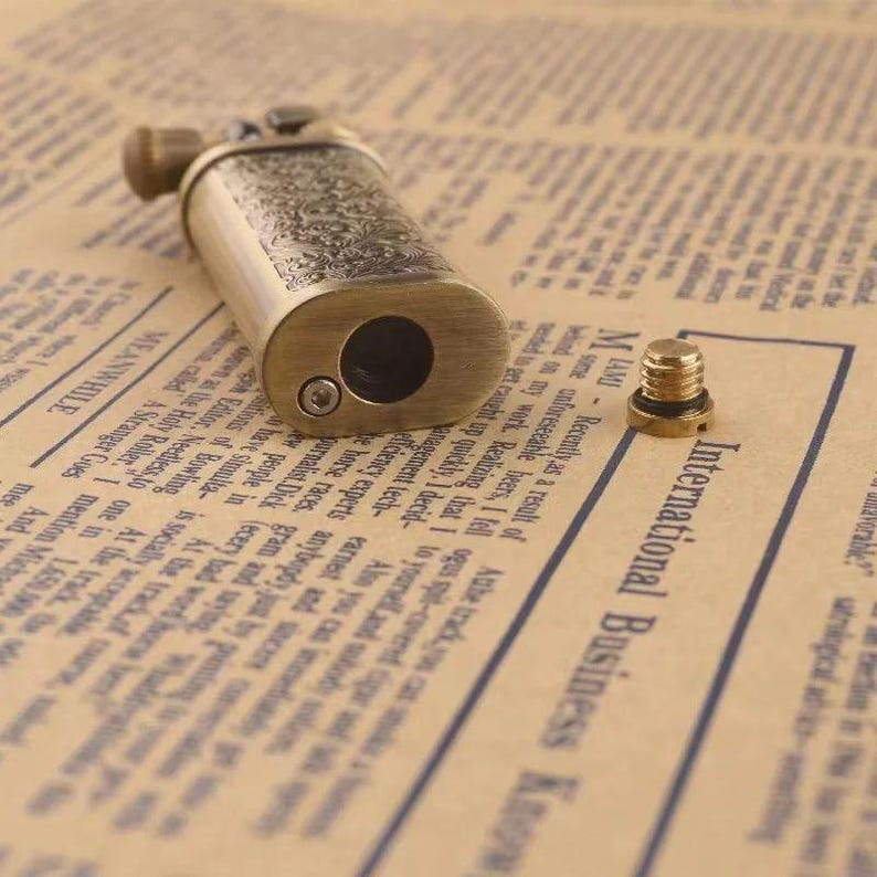 May include: A vintage-style brass lighter with intricate floral engravings. The lighter has a rectangular body with rounded edges and a large circular opening. A small, separate screw is also visible.