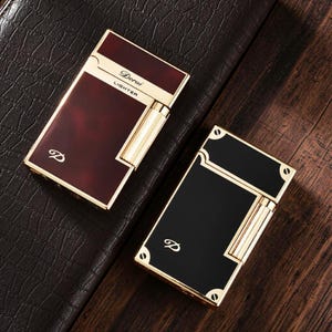 Puede incluir: Dos encendedores rectangulares con borde de metal dorado. El encendedor de la izquierda presenta un acabado jaspeado rojo oscuro con el texto Derui LIGHTER y un logotipo D. El de la derecha tiene un acabado negro sólido con cuatro tornillos.