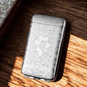 May include: A silver rectangular case with a hinged lid, featuring an intricate engraved pattern of leaves and a central crest with stylized initials. The case is resting on a wooden surface, with light reflecting off the metallic surface.