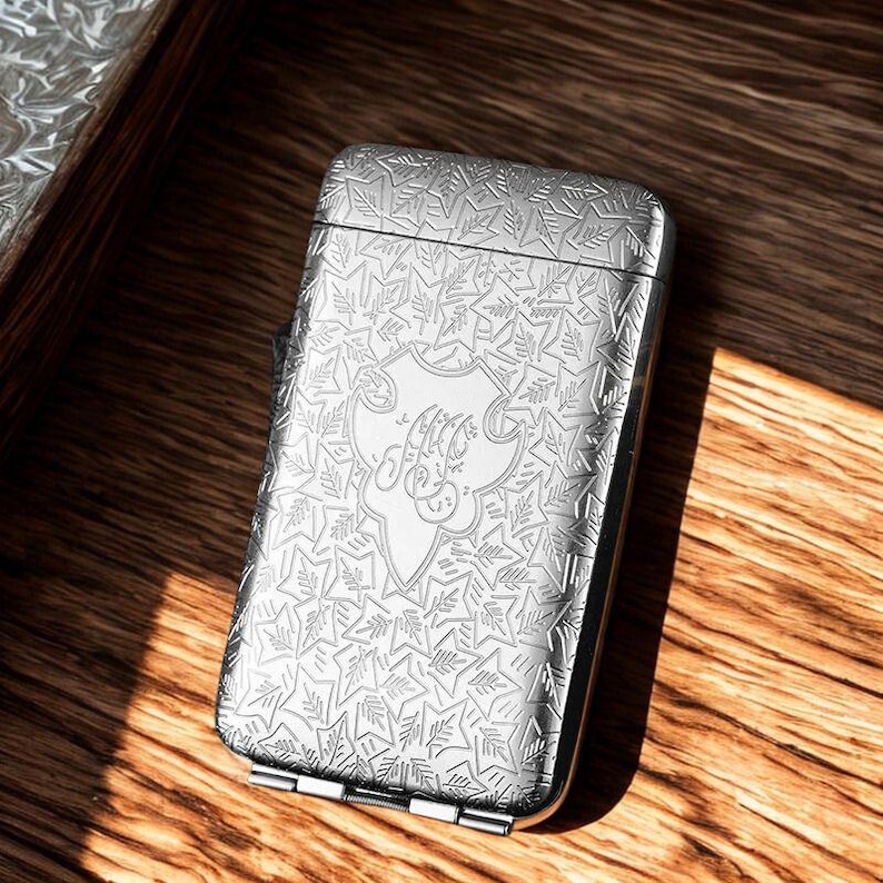 May include: A silver rectangular case with a hinged lid, featuring an intricate engraved pattern of leaves and a central crest with stylized initials. The case is resting on a wooden surface, with light reflecting off the metallic surface.