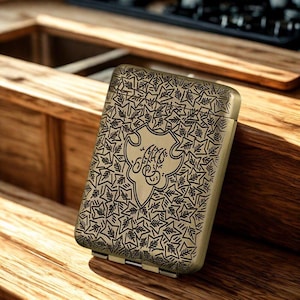 May include: An antique brass cigarette case with a detailed leaf pattern and a central emblem with stylized script. The hinged case is rectangular and appears to be made of metal.
