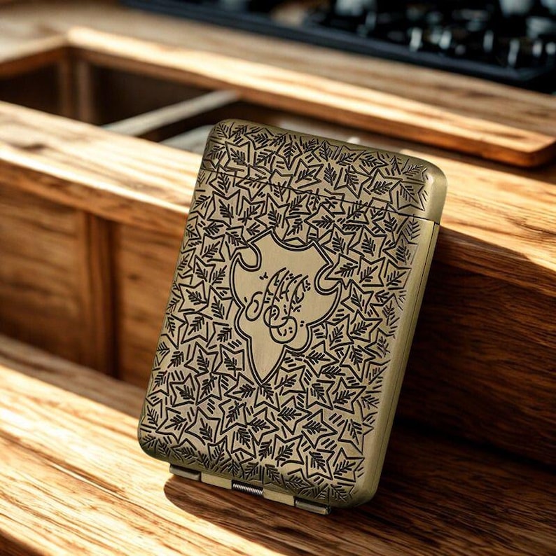 May include: An antique brass cigarette case with a detailed leaf pattern and a central emblem with stylized script. The hinged case is rectangular and appears to be made of metal.