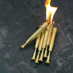 May include: A collection of brass-colored metal objects, some with flames emanating from the top. The objects have a cylindrical design with detailed ridges and a small, round base. The background is a dark, textured surface.