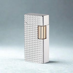 May include: A silver-colored lighter with a textured, diamond-patterned surface. The lighter has a rectangular shape with a gold-colored accent on the side. The top and side edges are smooth and shiny.