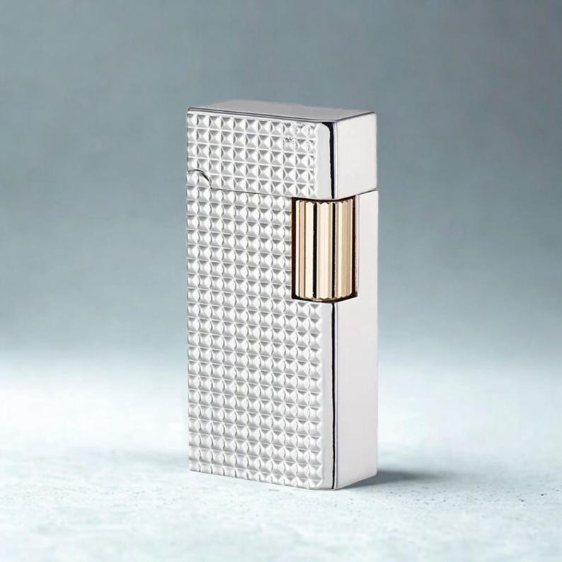 May include: A silver-colored lighter with a textured, diamond-patterned surface. The lighter has a rectangular shape with a gold-colored accent on the side. The top and side edges are smooth and shiny.