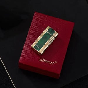 May include: A gold-toned lighter with a rectangular green panel, resting on a red box. The lighter has gold accents and the word "Derui" is printed on the lighter and the box. The box is a deep red color.