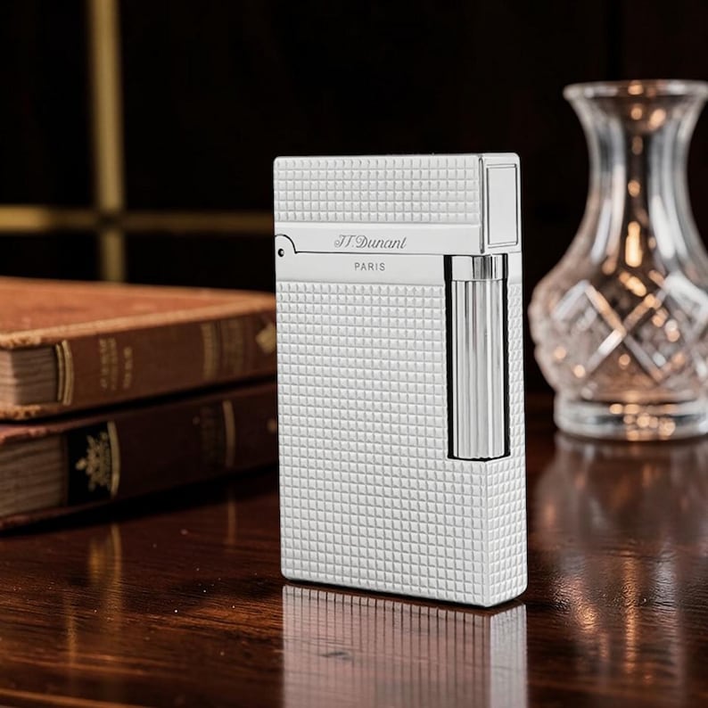 Puede incluir: Un encendedor plateado con un patr&oacute;n cuadrado texturizado. El encendedor tiene forma rectangular con una tapa con bisagras y una c&aacute;mara de combustible cil&iacute;ndrica. La marca "S.T. Dupont PARIS" es visible. El encendedor est&aacute; sobre una superficie de madera.