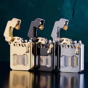Op de afbeelding: Drie metalen aanstekers in goud, zwart en zilver. Elke aansteker heeft een rechthoekige behuizing met een scharnierend deksel en een goudkleurige spuitmond. De aanstekers worden gepresenteerd tegen een donkerblauwe achtergrond.