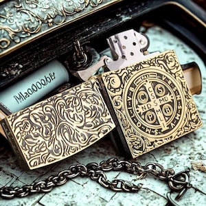 May include: A gold-colored metal lighter with intricate engraved patterns and a chain. The lighter is open, revealing the interior. The design includes a cross and the letters "CSMD" and "PAX".