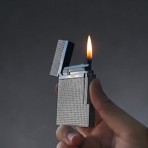 Puede incluir: Un encendedor plateado con una superficie texturizada con patr&oacute;n de diamantes y una tapa abierta, emitiendo una llama naranja brillante. La palabra "PARIS" es visible en el encendedor. El encendedor se sostiene en una mano sobre un fondo gris oscuro.