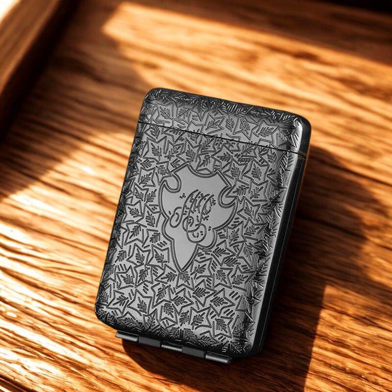 May include: A dark grey rectangular case with an engraved floral pattern. The case features a central shield design with stylized script. The case is closed and has a hinged bottom.