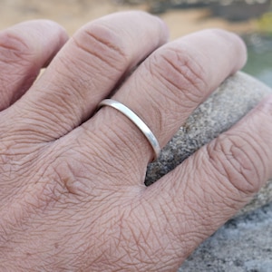 Puede incluir: Un anillo fino de plata se lleva en un dedo. El anillo es una banda sencilla con un acabado pulido. El tono de piel de la mano es claro y el fondo está borroso.