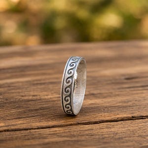 Puede incluir: Un anillo de plata con un patrón de ondas repetidas alrededor de la banda. El anillo está colocado sobre una superficie de madera con un fondo borroso. El diseño del anillo presenta una serie de formas arremolinadas conectadas, creando un patrón continuo.