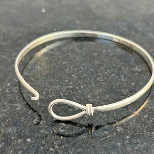 May include: A silver bangle bracelet with a loop and clasp closure. The bracelet is made of a thin, round metal band. The loop closure is secured with a wrapped wire design. The bracelet is shown on a dark, textured surface.