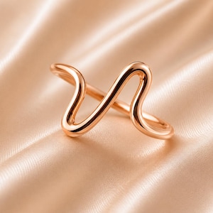 Può includere: Un anello in oro rosa dal design unico e fluido. L'anello presenta una forma ondulata continua che avvolge il dito. La superficie liscia e lucida riflette la luce, creando una sottile lucentezza. L'anello è posto su uno sfondo satinato.