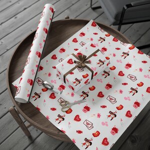May include: White wrapping paper with a repeating pattern of red hearts, cupcakes, envelopes, and figures. A wrapped gift with a brown ribbon sits on the paper, along with a roll of the same paper and scissors.