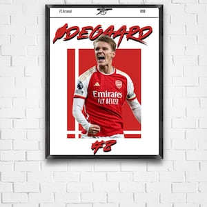 May include: Framed poster featuring a soccer player in a red and white jersey with the text "ØDEGAARD" in a stylized font. The poster includes the text "FC Arsenal" and "1998". The player's jersey displays the "Emirates FLY BETTER" logo and the number 8.
