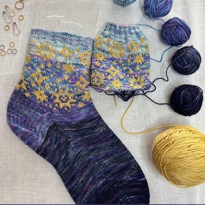 Pode incluir: Meias de malha feitas à mão com um padrão de estrelas em tons de azul, roxo e amarelo. A imagem inclui novelos de lã e o texto "CHARMED STARS SOCKS". As meias são rotuladas como "SMALL | MEDIUM | LARGE".