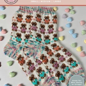 May include: A pair of knitted baby socks with a repeating pattern of brown teddy bears wearing teal, pink, and orange outfits on a white background. The socks are labeled "Baby Bear Socks" and are available in small, medium, and large sizes. The socks are surrounded by heart-shaped candies.