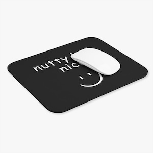 May include: Black rectangular mousepad with the words "nutty nice" and a smiling face in white. A white wireless mouse is on the mousepad. The mousepad is designed for computer use.