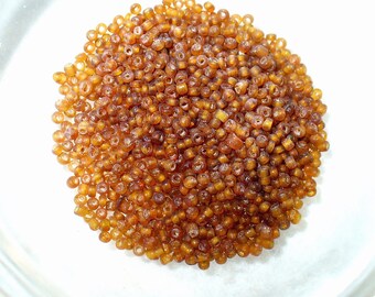 May include: A pile of small, amber-colored seed beads. The beads are round with a hole through the center, and they are scattered on a white surface. The beads have a glossy finish and are likely made of glass or plastic.