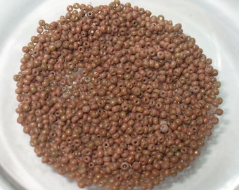 May include: A close-up shot of a pile of small, round, brown beads. The beads are a muted, earthy tone, and appear to be made of glass or a similar material. They are scattered in a clear, shallow bowl, suggesting they are for crafting or jewelry making.