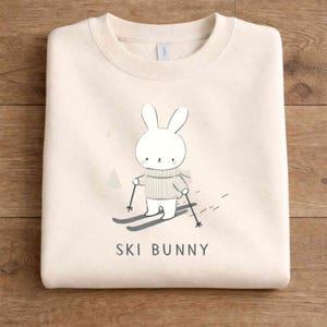 May include: Cream-colored sweatshirt featuring a cartoon bunny skiing. The bunny wears a gray sweater and scarf, holding ski poles. The words "SKI BUNNY" are printed below the illustration. The sweatshirt is folded on a wooden surface.