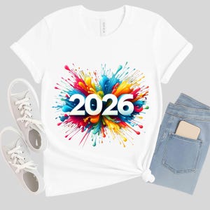 Peut inclure: T-shirt blanc avec un motif éclaboussures coloré affichant l'année "2026" en lettres blanches. Le motif comprend des teintes vives de rouge, bleu, jaune et vert. Le t-shirt est associé à un jean bleu clair et des baskets blanches.